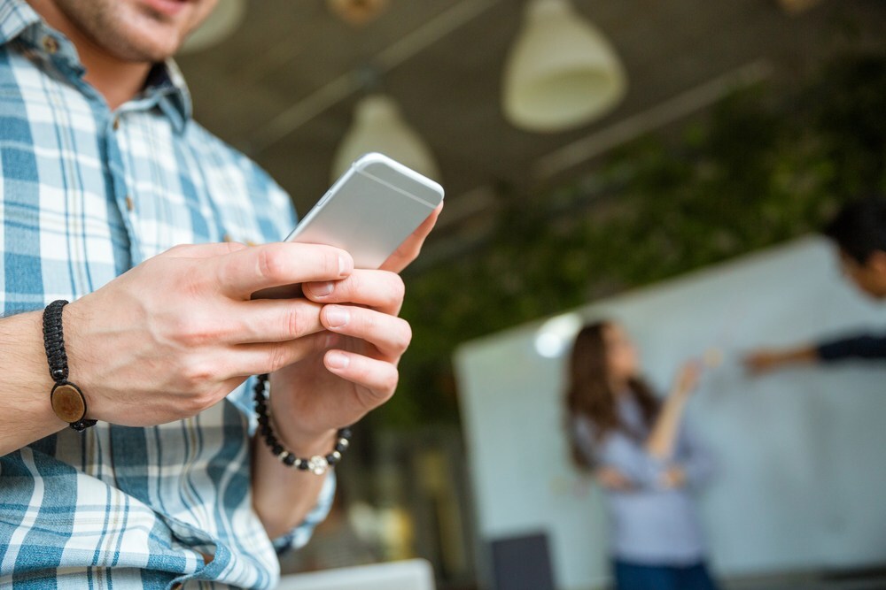 Closeup of hands of young man in checkered shirt using mobile phone while his partners arguing_RD