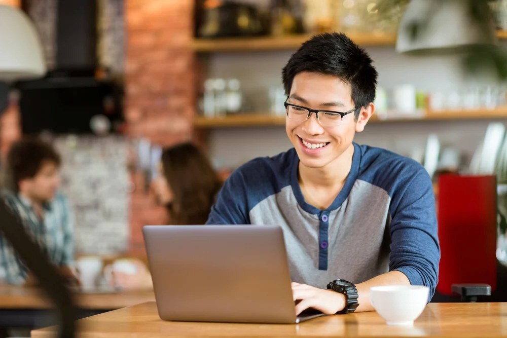 Happy cheerful young asian male in glasses smiling and using laptop in cafe_RD